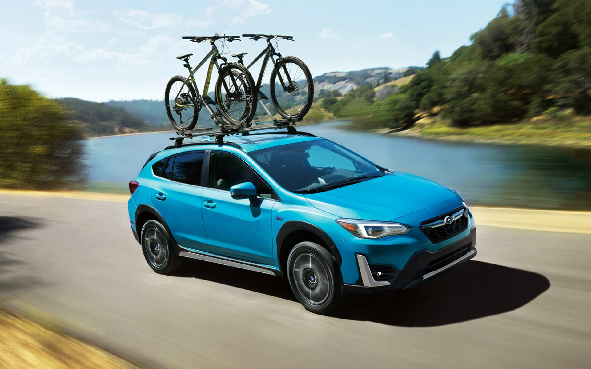 A blue Crosstrek Hybrid with two bicycles on its roof rack driving beside a river | Zappone Subaru Norwich in Norwich NY