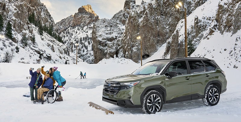 A Subaru Forester Hybrid parked on a snowy mountain road near a group of people gathered around a wheelchair user, with snow-covered cliffs and pine trees in the background under a partly cloudy sky.