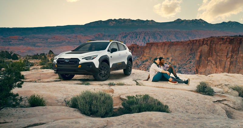 A static image of a white 2026 Subaru Wilderness model parked on a flat rock surface overlooking a wide desert canyon.